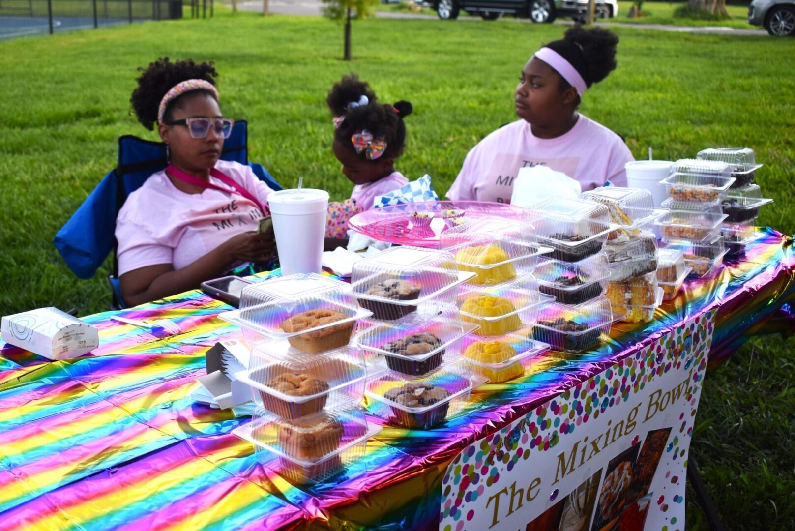 Three girls selling baked goods at a colorful outdoor market stall.