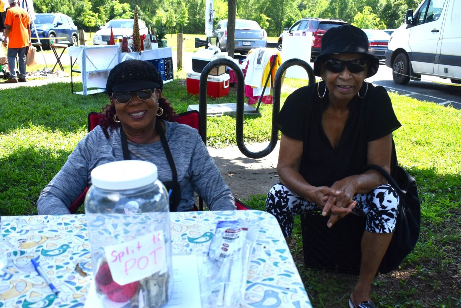 Two women sitting outdoors at a table with a jar labeled for donations.