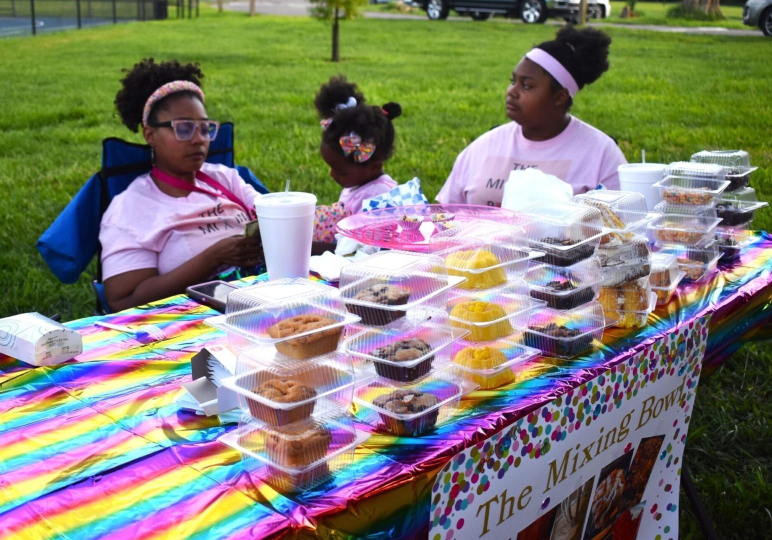 Three girls selling baked goods at a colorful outdoor market stall.