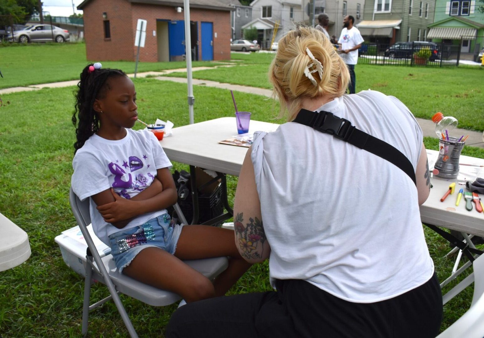 A young girl and woman interact at an outdoor table under a tent.